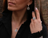 A woman wearing a sterling silver jewelry set consisting of a ring, necklace, and earrings with a geometric design, standing outdoors.