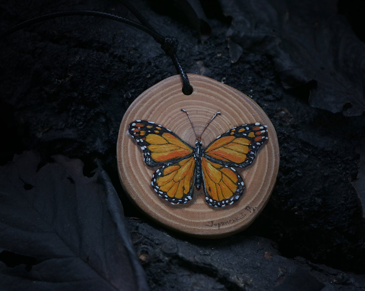 May include: A wooden pendant with a hand-painted monarch butterfly design. The pendant is round and has a black cord.