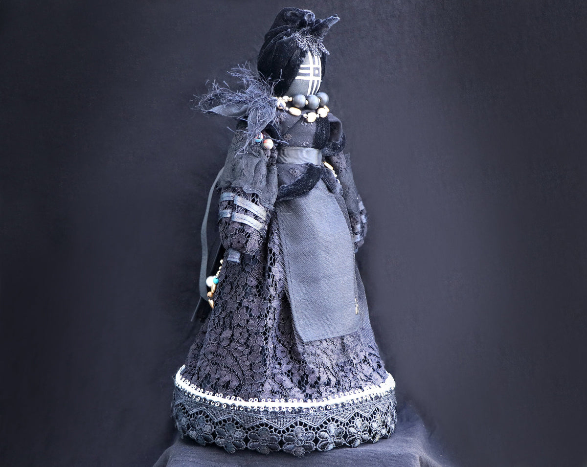May include: A black fabric doll wearing a black lace dress with a white trim and a black velvet hat. The doll has a black face with white markings and is wearing a beaded necklace.