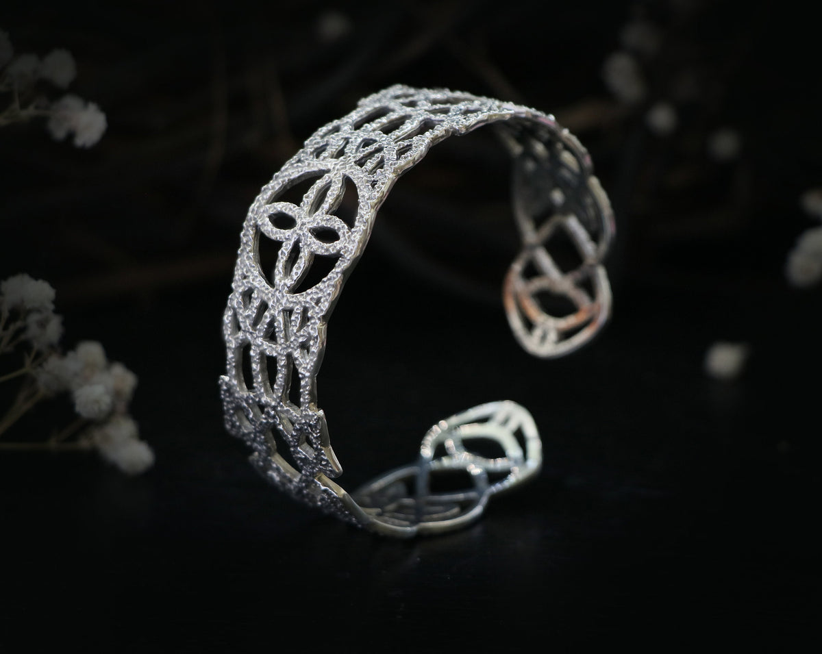 May include: A silver bracelet with a delicate, intricate design. The bracelet is made of a thin metal and has a rounded shape.
