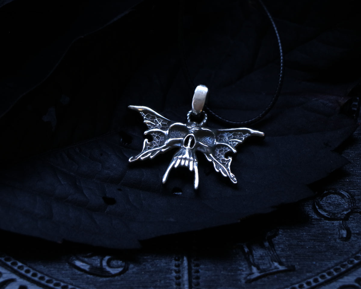 A silver Luna Moth necklace, pendant in the shape of a skull moth against a dark background, displayed on a circular object with letters.