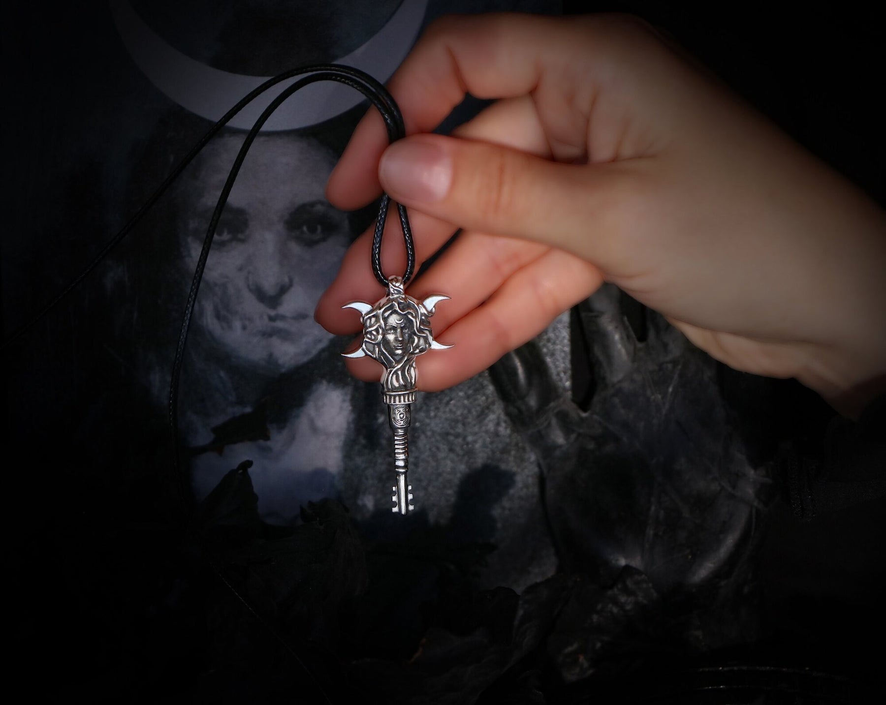 A hand holding a gothic-style silver Hecate key pendant on a black cord necklace, set against a dark, moody background featuring a skull and a moon-phase print.