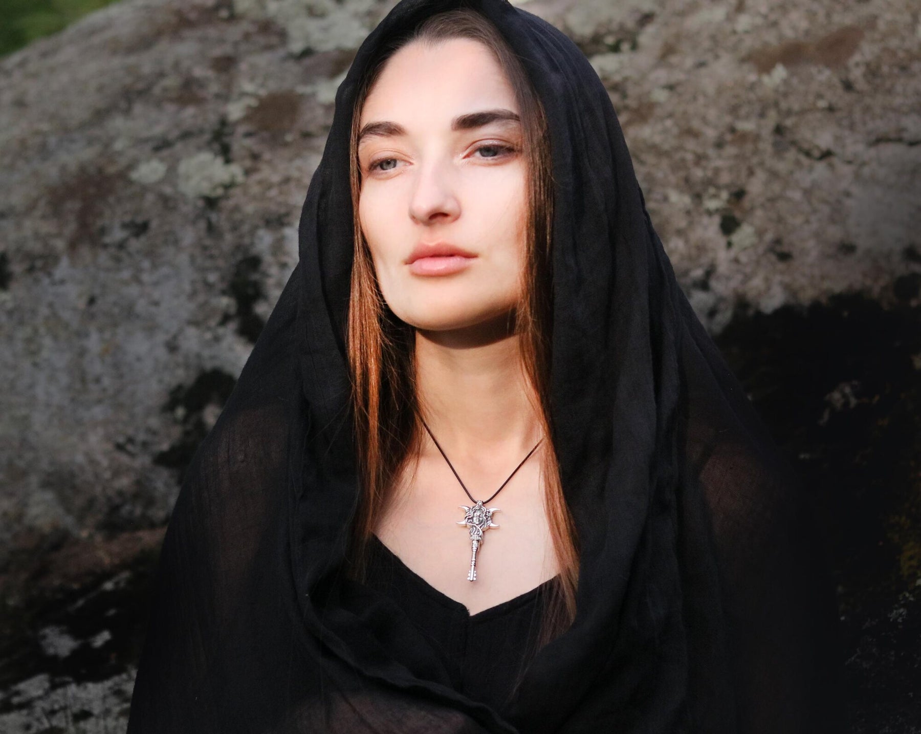 A woman wearing a black sheer head covering and black top, with a silver Hecate Key pendant necklace, posed outdoors against a large rock.