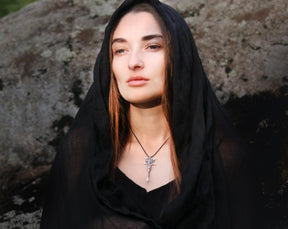 A woman wearing a black sheer head covering and black top, with a silver Hecate Key pendant necklace, posed outdoors against a large rock.