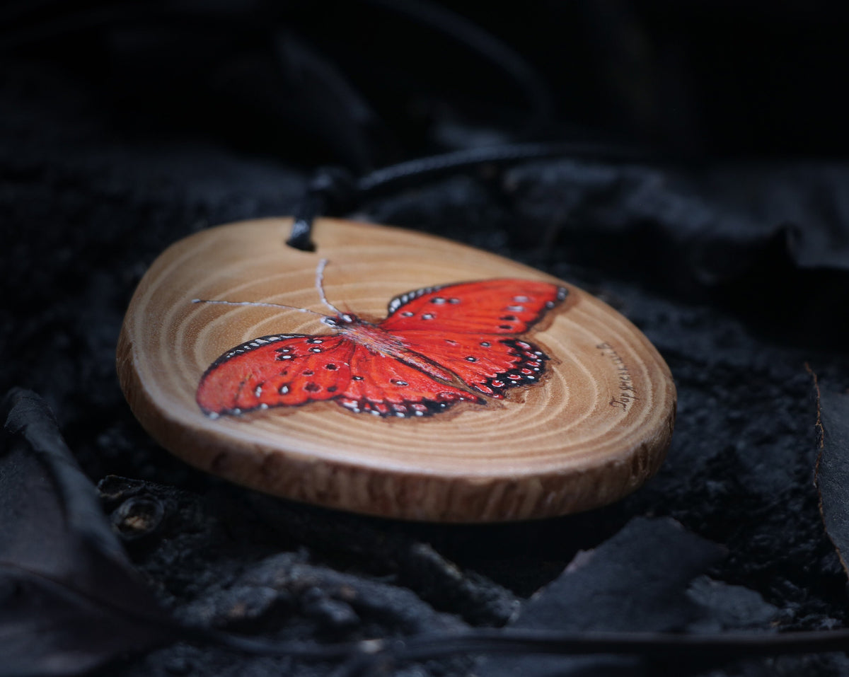 May include: A wooden pendant with a hand-painted red butterfly. The pendant is round and has a natural wood grain. The butterfly has black markings and is set against a light brown background.