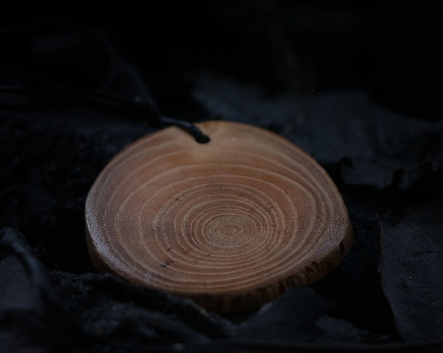 May include: A wooden pendant with a circular shape and a dark brown cord. The pendant has a natural wood grain pattern with concentric circles.