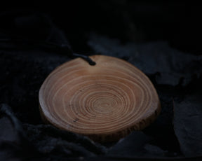 May include: A wooden pendant with a circular shape and a dark brown cord. The pendant has a natural wood grain pattern with concentric circles.