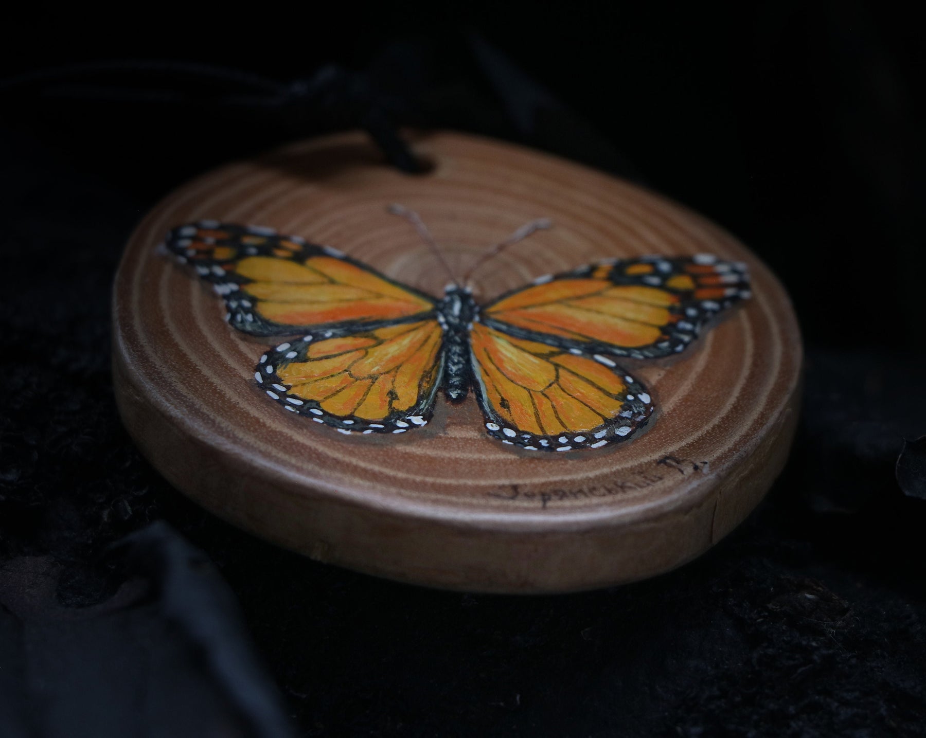May include: A wooden round ornament with a painted orange and black butterfly. The ornament has a brown wood grain and a black cord for hanging.