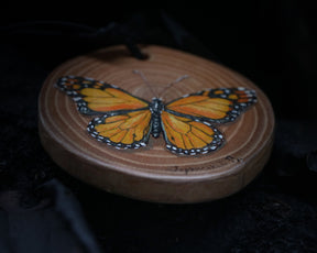 May include: A wooden round ornament with a painted orange and black butterfly. The ornament has a brown wood grain and a black cord for hanging.