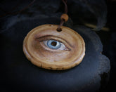 May include: A wooden pendant with a painted eye. The eye is blue with brown eyelashes and a light brown eyebrow. The pendant is attached to a brown leather cord.