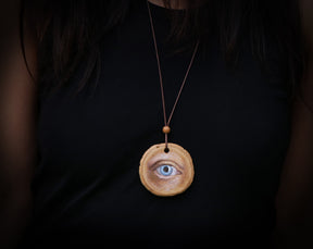 May include: A wooden pendant necklace with a painted blue eye on a brown wood background.