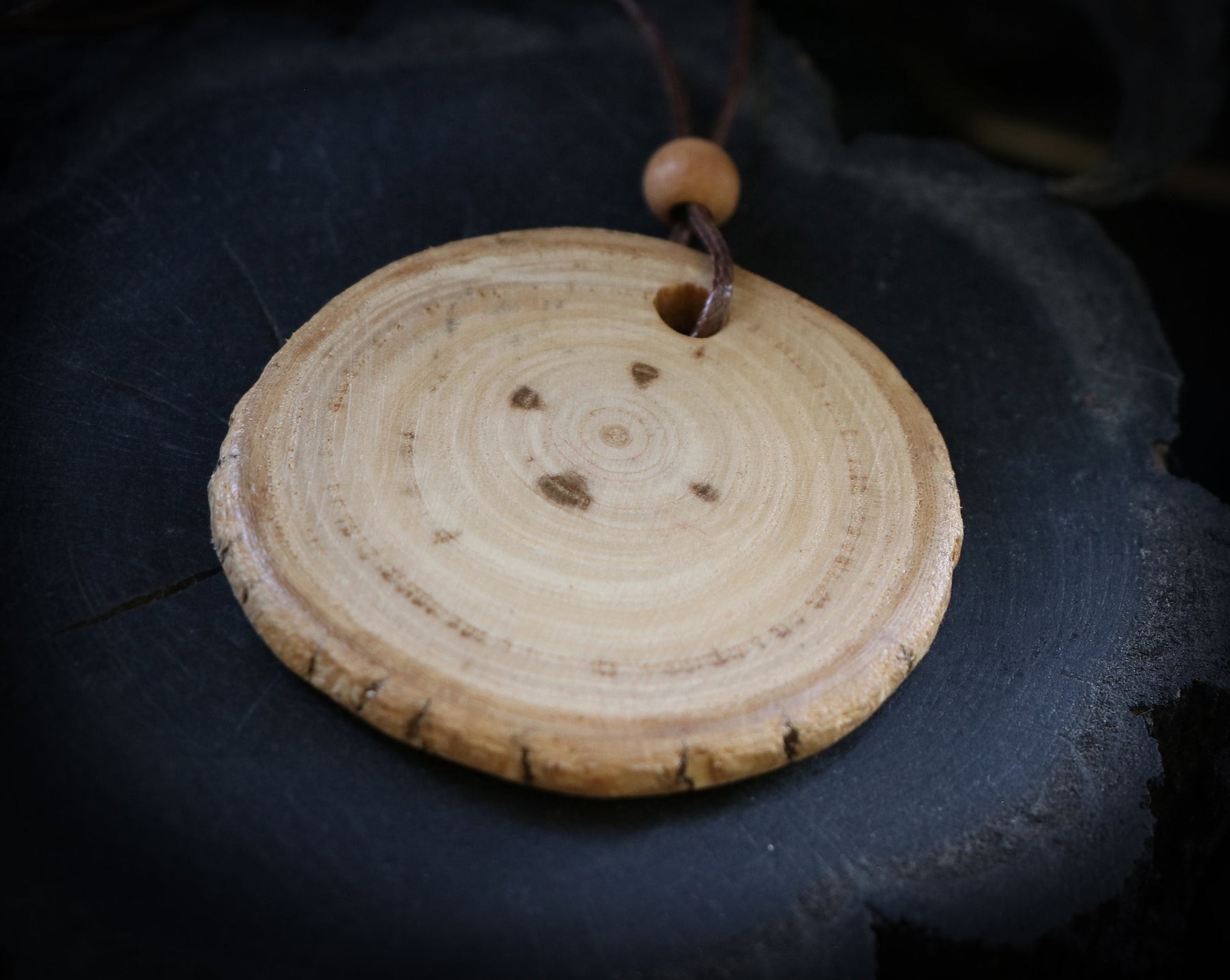 May include: A round wooden pendant with a natural wood finish and a brown leather cord. The pendant has a circular shape with concentric rings and a hole in the center.