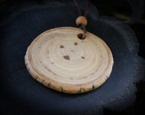 May include: A round wooden pendant with a natural wood finish and a brown leather cord. The pendant has a circular shape with concentric rings and a hole in the center.