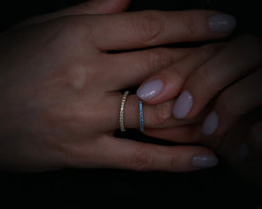 May include: Two thin diamond eternity bands on a finger. The top band is set with small round diamonds and the bottom band is set with small blue diamonds.