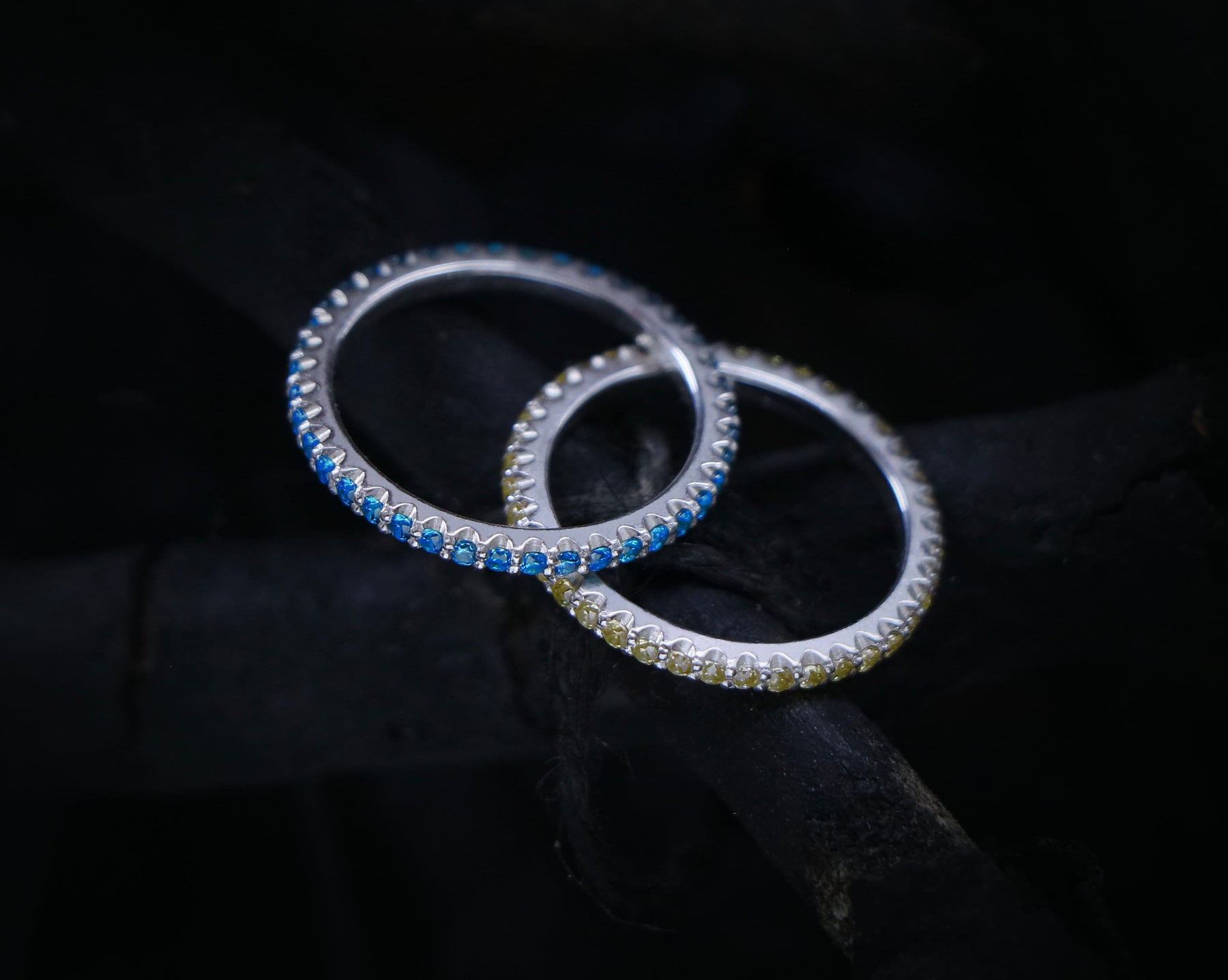 May include: Two silver rings with a row of blue and yellow gemstones. The rings are interlocked and set against a dark background.