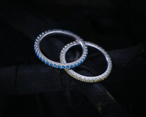 May include: Two silver rings with a row of blue and yellow gemstones. The rings are interlocked and set against a dark background.