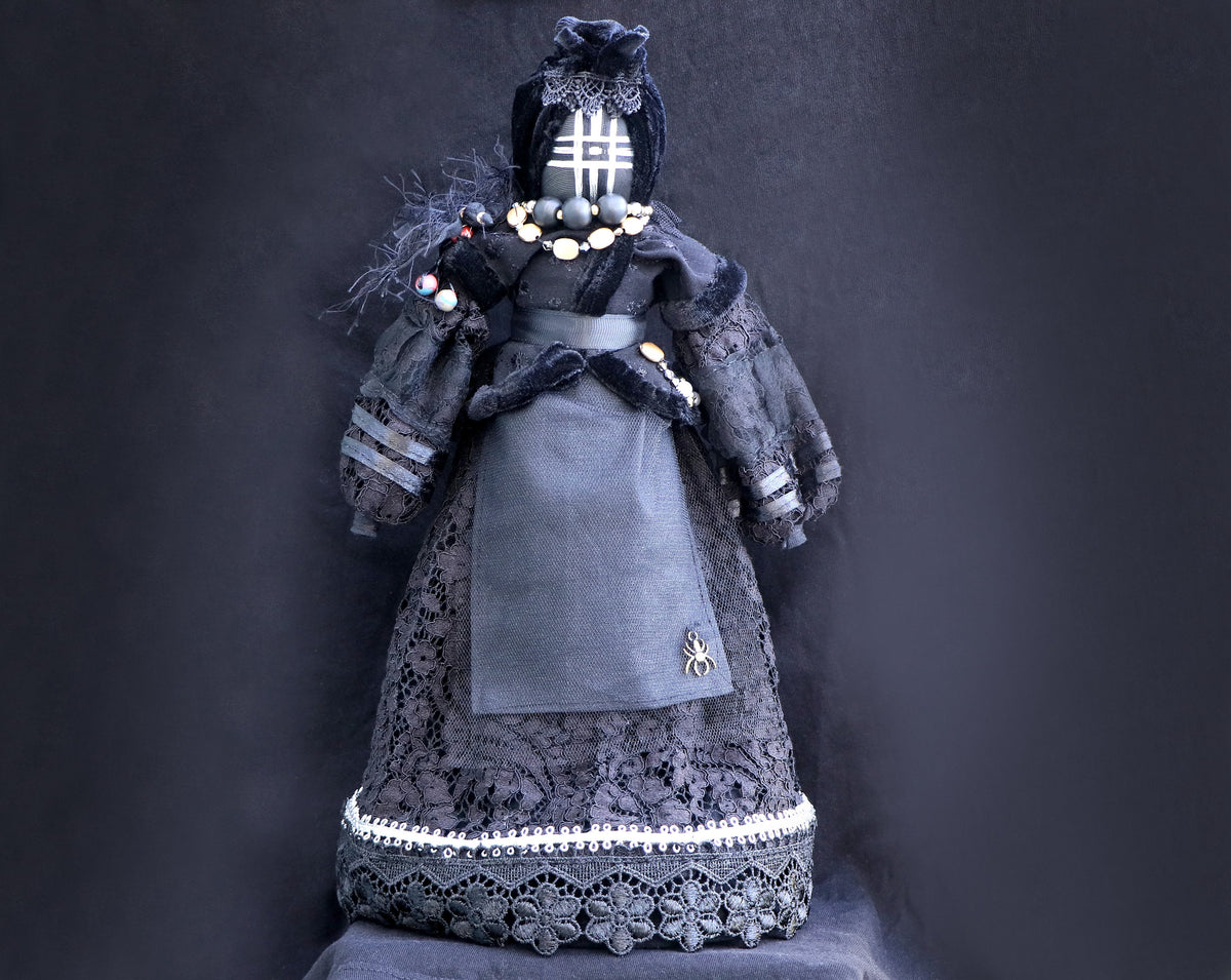 May include: A black doll wearing a black lace dress with a spider brooch. The doll has a black face with a white grid pattern and wears a black hat with a white lace trim.