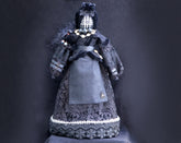 May include: A black doll wearing a black lace dress with a spider brooch. The doll has a black face with a white grid pattern and wears a black hat with a white lace trim.