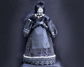May include: A black doll wearing a black lace dress with a spider brooch. The doll has a black face with a white grid pattern and wears a black hat with a white lace trim.