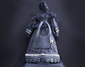 May include: A black doll wearing a black velvet hooded dress with lace trim and a black ribbon around the waist.