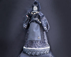 May include: A black fabric doll wearing a black lace dress and a black hooded cloak. The doll has a black and white patterned face and is adorned with beads and a spider brooch.