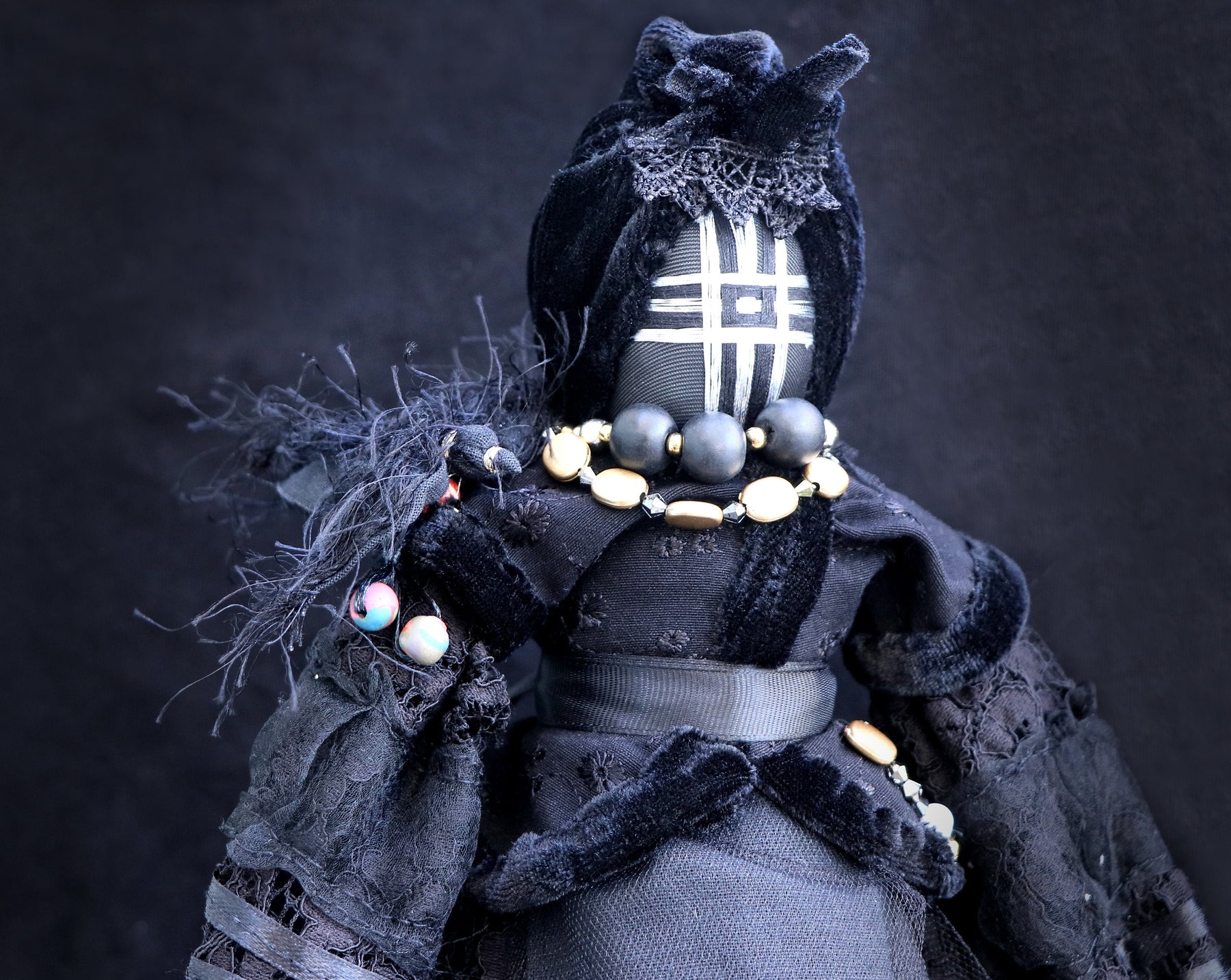 May include: A black fabric doll with a black velvet dress and a black and white patterned face. The doll is wearing a necklace of gold and black beads.