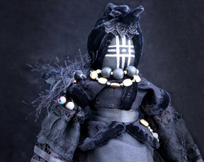 May include: A black fabric doll with a black velvet dress and a black and white patterned face. The doll is wearing a necklace of gold and black beads.
