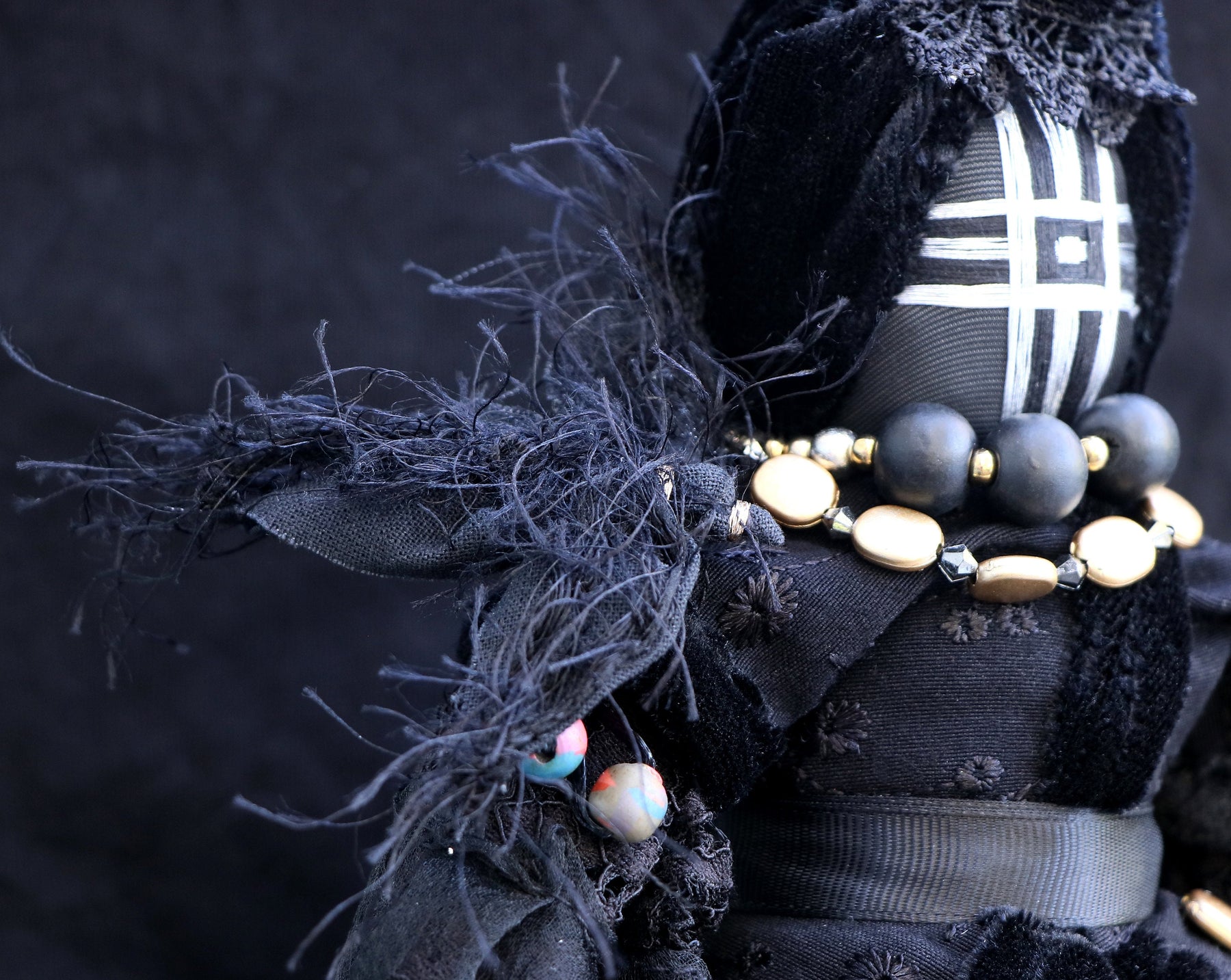 May include: A black fabric doll with a white geometric pattern on its face. The doll is wearing a black velvet dress with gold and black beads around its neck.