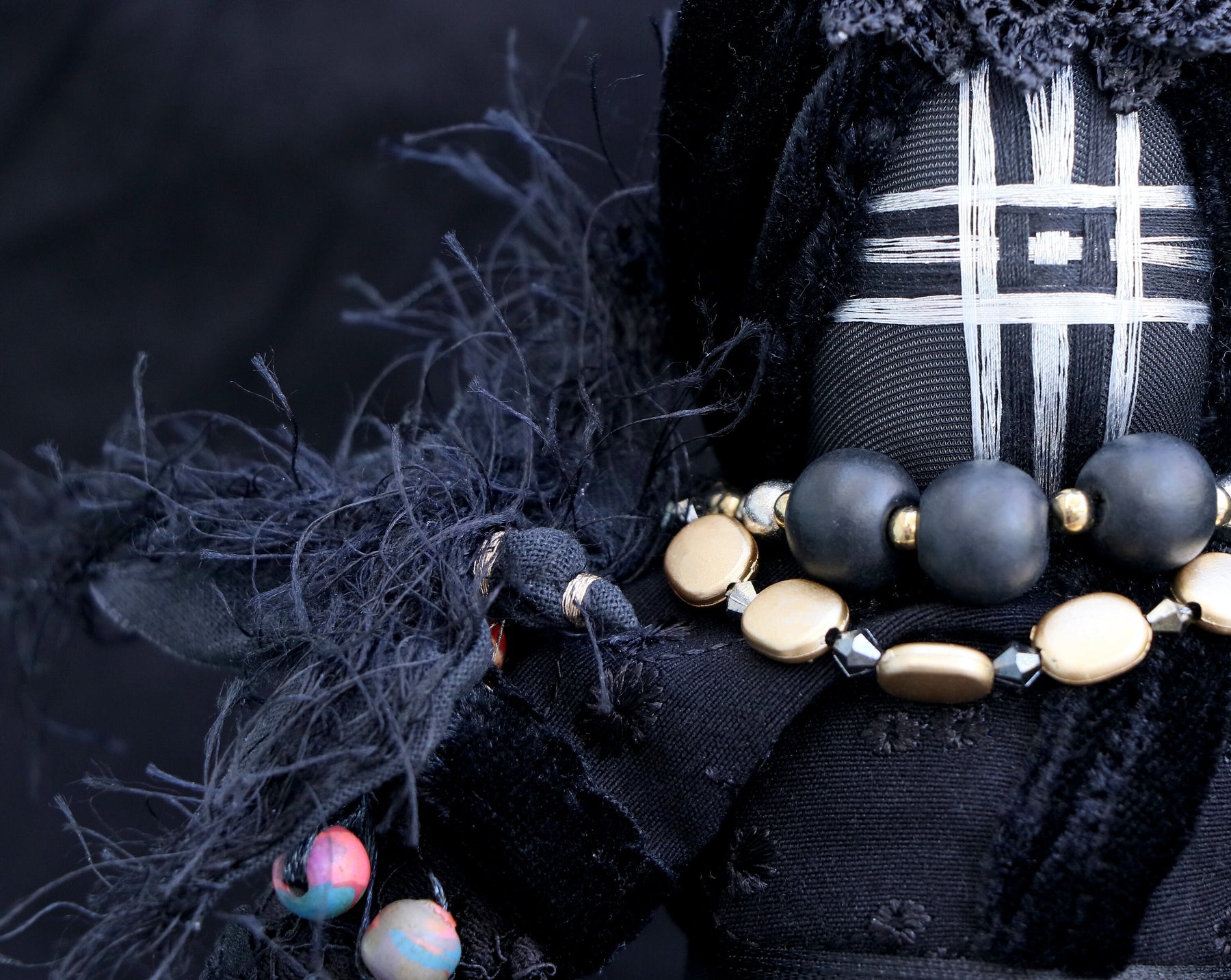 May include: Black fabric doll with a geometric pattern on the face, wearing a necklace of black and gold beads.