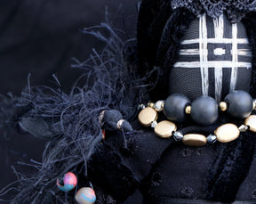 May include: Black fabric doll with a geometric pattern on the face, wearing a necklace of black and gold beads.