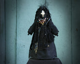 May include: Black fabric doll with long black hair, wearing a black lace dress and a beaded necklace with turquoise and gold accents. The doll has a black face with a white painted design and a spider pin on the dress.