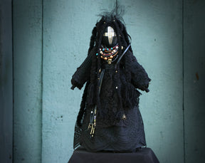 May include: Black fabric doll with long black hair, wearing a black lace dress and a beaded necklace with turquoise and gold accents. The doll has a black face with a white painted design and a spider pin on the dress.