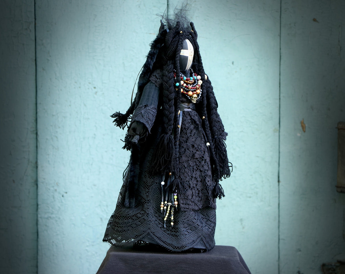 May include: A black fabric doll with long black hair and a white cross on its face. The doll is wearing a black lace dress and a necklace with colorful beads.