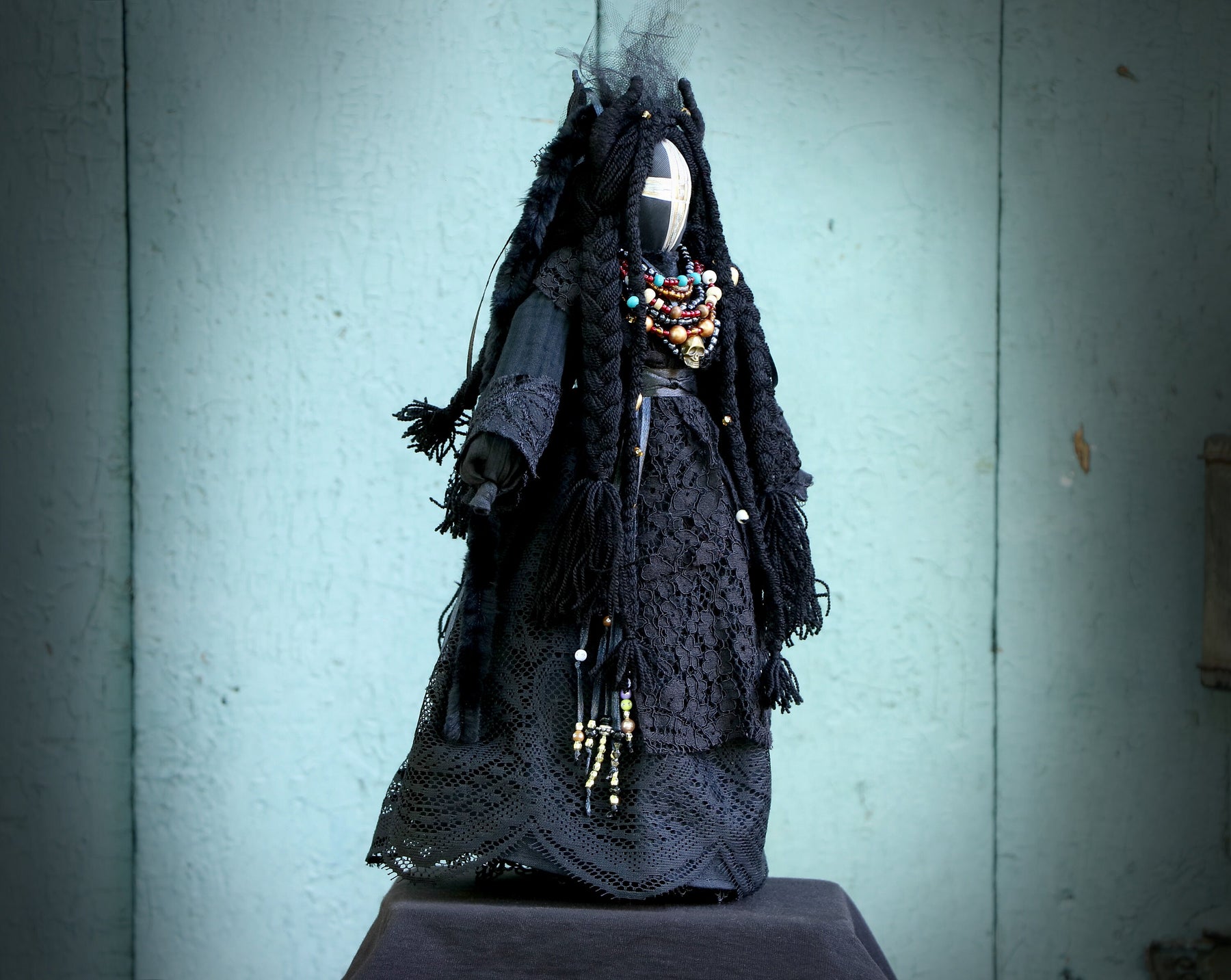 May include: A black fabric doll with long black hair and a white cross on its face. The doll is wearing a black lace dress and a necklace with colorful beads.