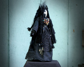 May include: A black fabric doll with long black hair and a white cross on its face. The doll is wearing a black lace dress and a necklace with colorful beads.