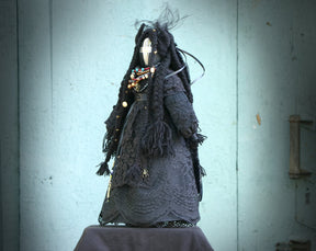 May include: A black fabric doll with long black hair and a white face. The doll is wearing a black lace dress and has a variety of beads and charms around its neck.