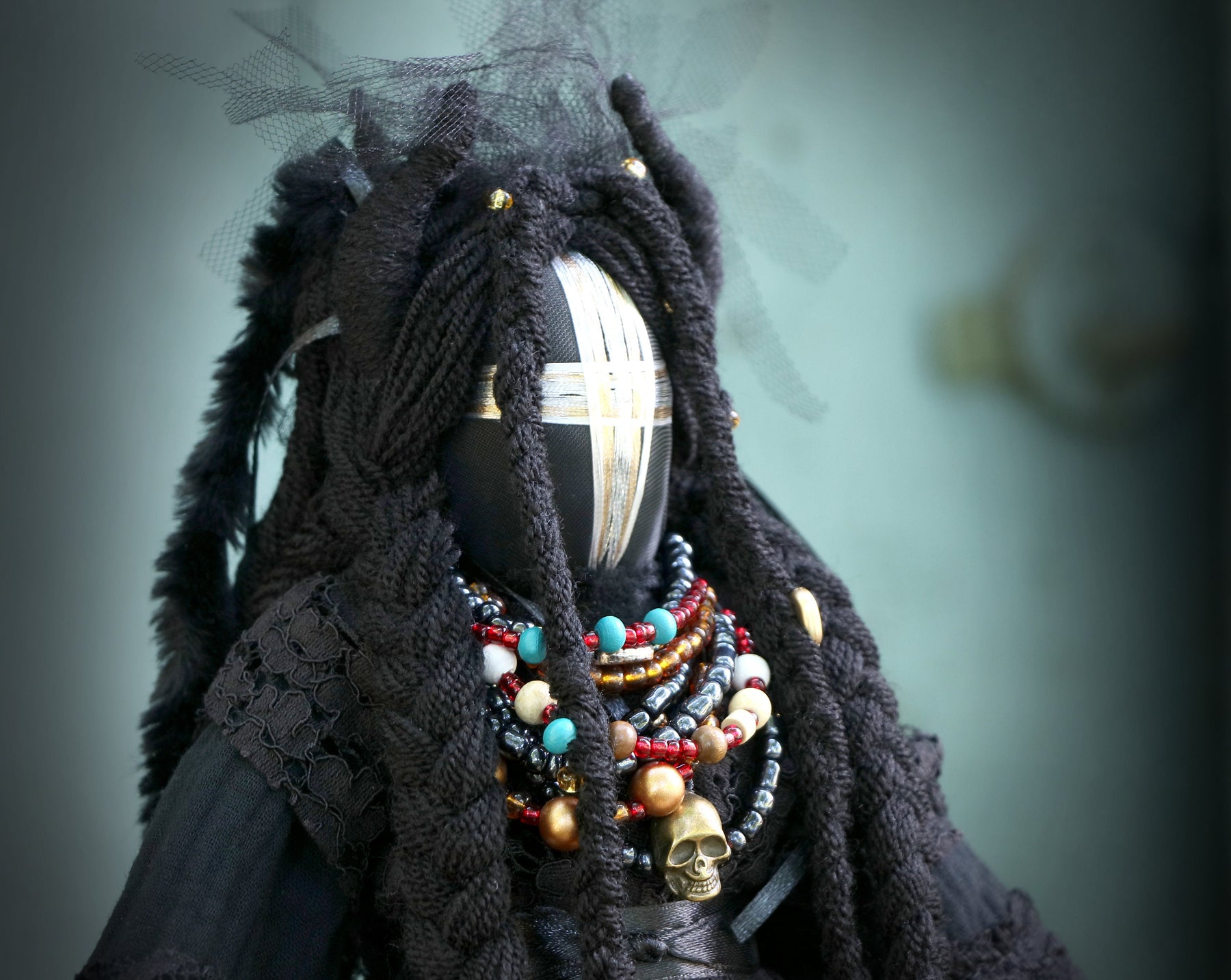 May include: A black doll with long, braided hair and a silver and black face mask. The doll is wearing a black lace dress and a variety of colorful beaded necklaces.