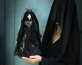 May include: A woman wearing a black veil holds a black doll with a white cross on its face. The doll is adorned with beads, charms, and lace.