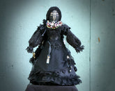 May include: A black fabric doll wearing a black lace dress with a beaded necklace and a black fabric head covering with a cross design.