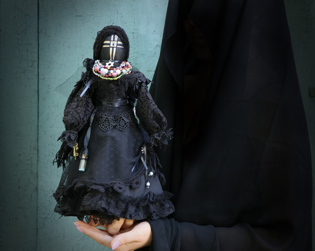 May include: A black doll with a white cross on its face, wearing a black lace dress and a beaded necklace. The doll is being held by a person wearing a black robe.