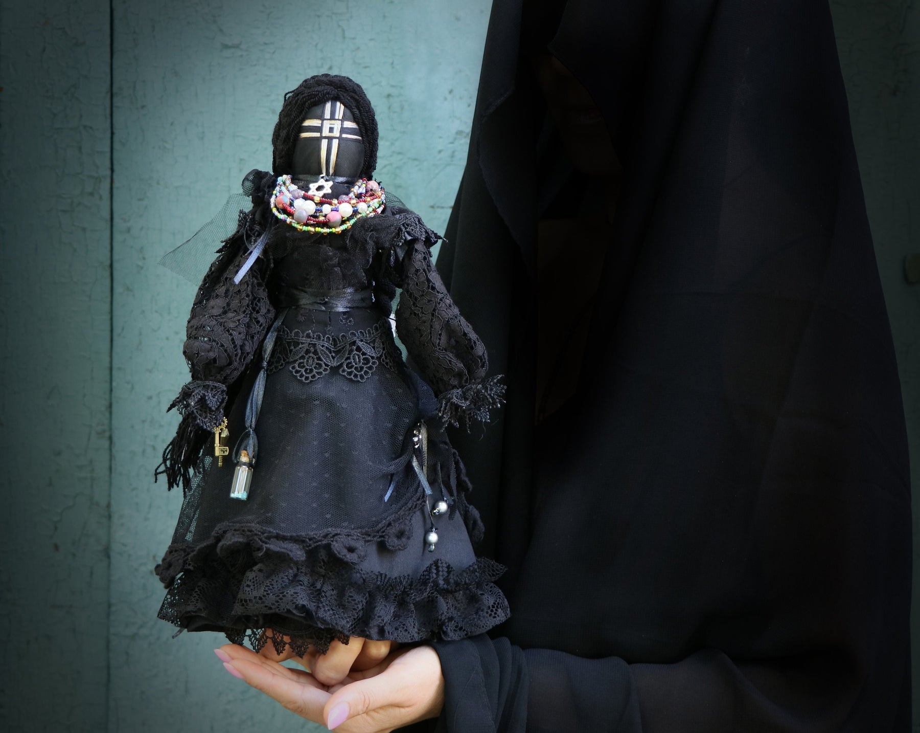 May include: A black doll with a white cross on its face, wearing a black lace dress and a beaded necklace. The doll is being held by a person wearing a black robe.