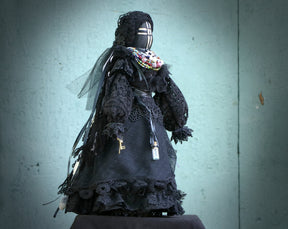 May include: A black fabric doll wearing a black lace dress and a veil. The doll has a black and white face with a cross design and is adorned with beaded necklaces.