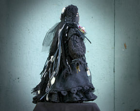 May include: A black doll wearing a long black dress with lace trim and a black veil. The doll has a black face and is wearing a black hat with white accents. The doll is standing on a dark surface.