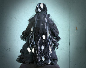 May include: A black and white doll wearing a black lace dress with white accents. The doll is dressed in a gothic style and has a veil over its head.