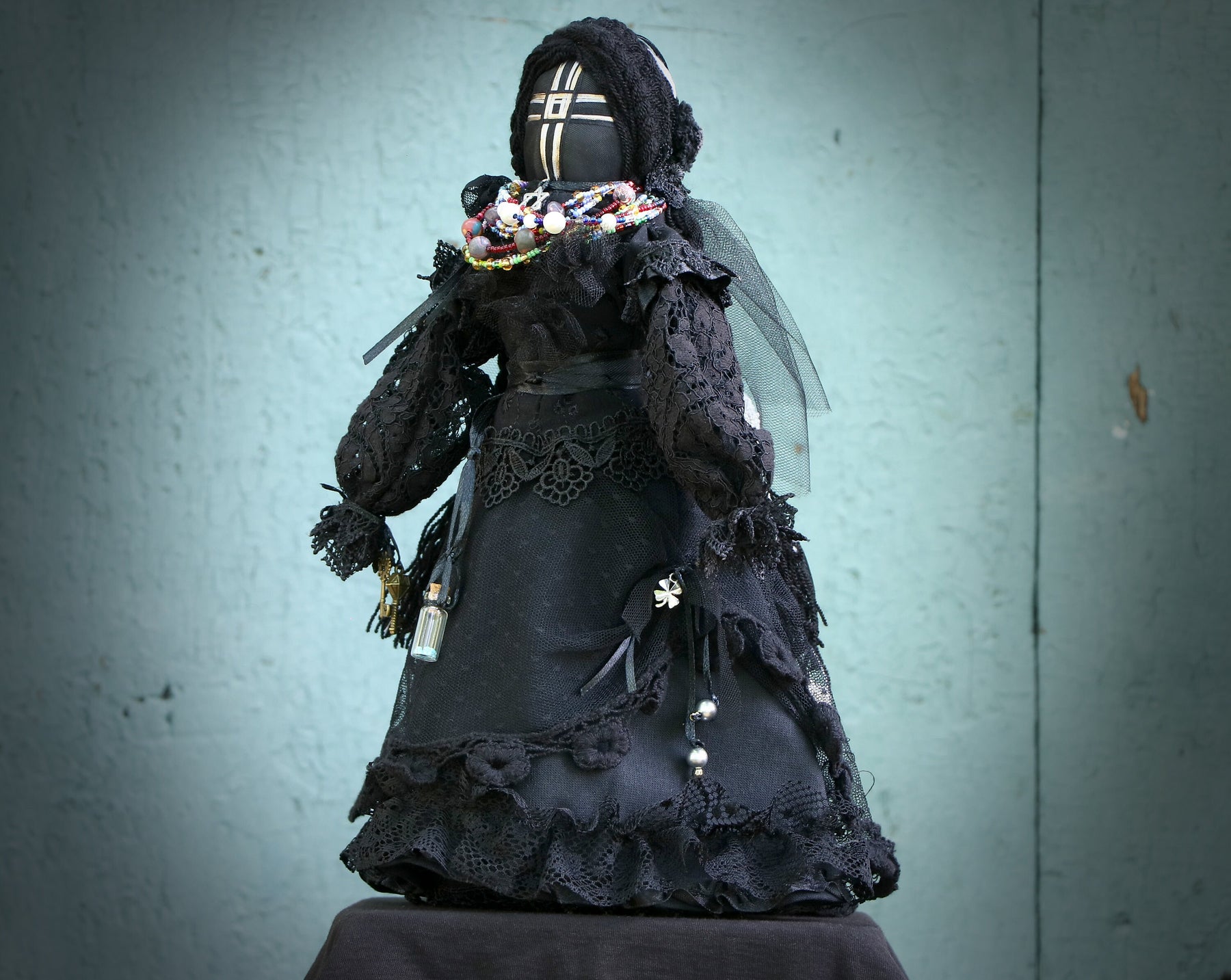 May include: A black fabric doll wearing a black lace dress with a veil and a beaded necklace. The doll has a black fabric head with a white cross design.