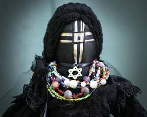 May include: A black fabric doll with a black and gold cross on its face. The doll is wearing a black lace dress and a necklace with colorful beads and a silver star pendant.