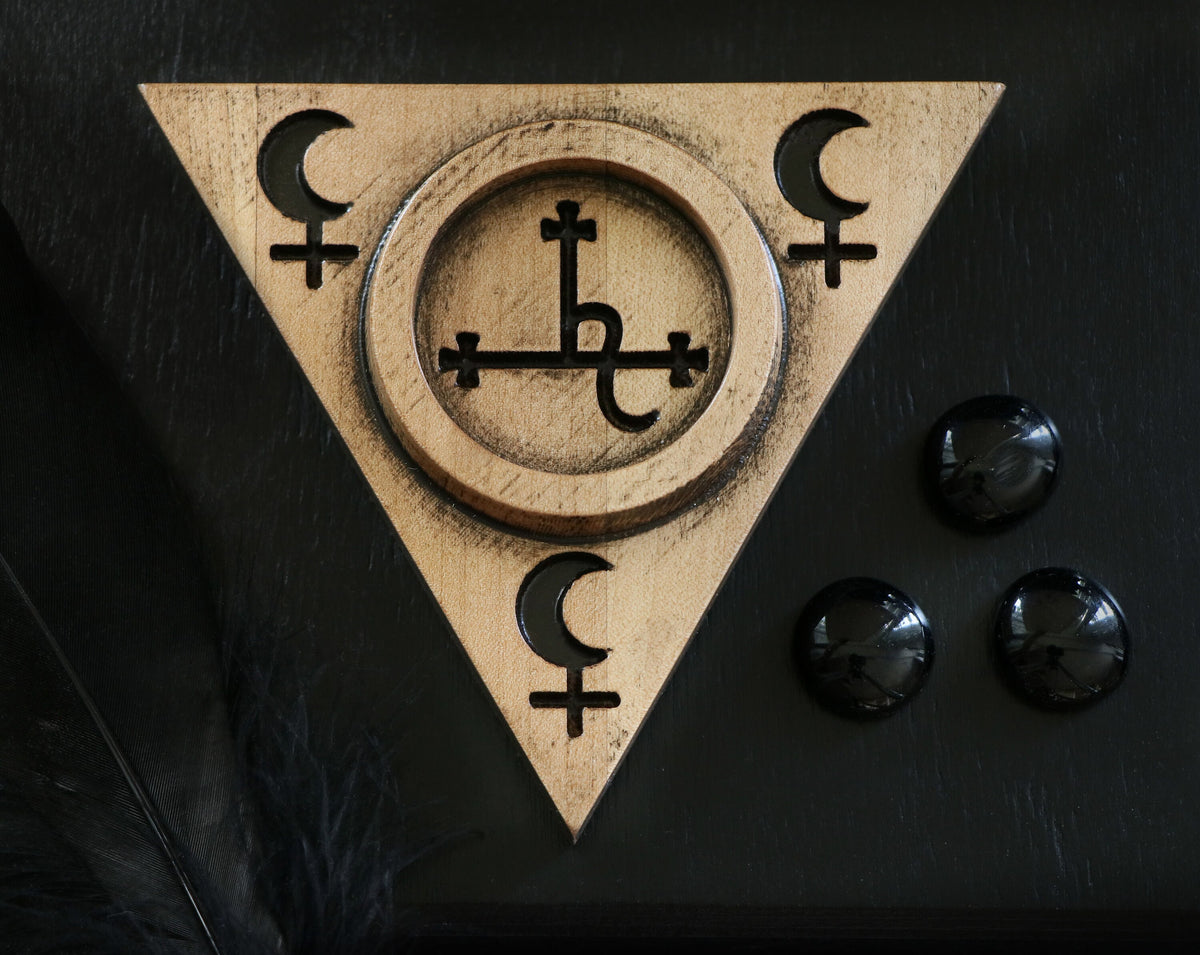 May include: A gold-toned wooden triangle with a circle in the center. The circle has a cross with a symbol inside. The triangle has three crescent moons and crosses on each point. The triangle is on a black surface with three black round stones.