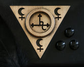 May include: A gold-toned wooden triangle with a circle in the center. The circle has a cross with a symbol inside. The triangle has three crescent moons and crosses on each point. The triangle is on a black surface with three black round stones.