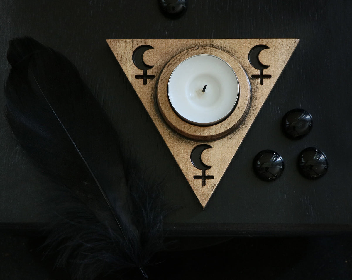 May include: A wooden triangle candle holder with a white tea light candle in the center. The triangle has a crescent moon and a female symbol on each side. The holder is on a black surface with black stones.