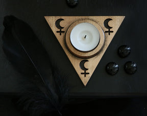 May include: A wooden triangle candle holder with a white tea light candle in the center. The triangle has a crescent moon and a female symbol on each side. The holder is on a black surface with black stones.